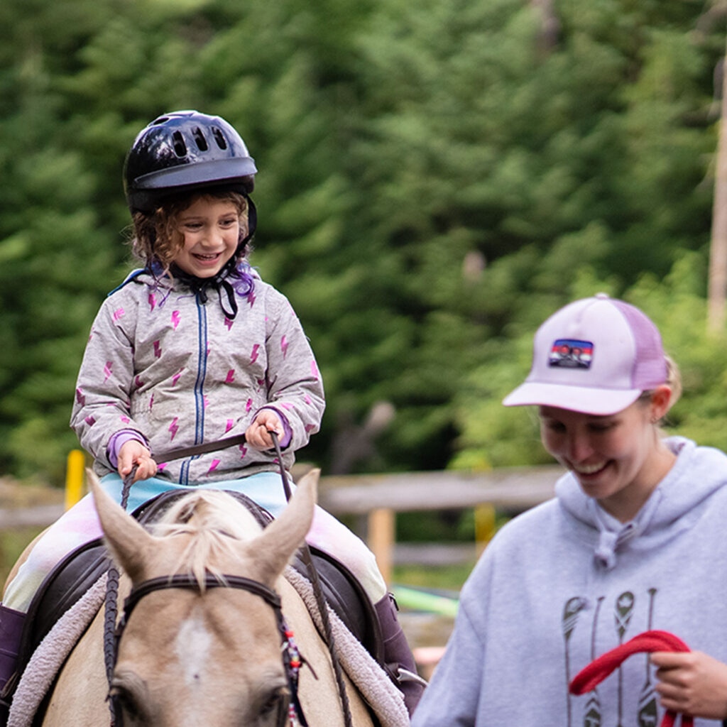 small child smiling on horseback with riding teacher assisting