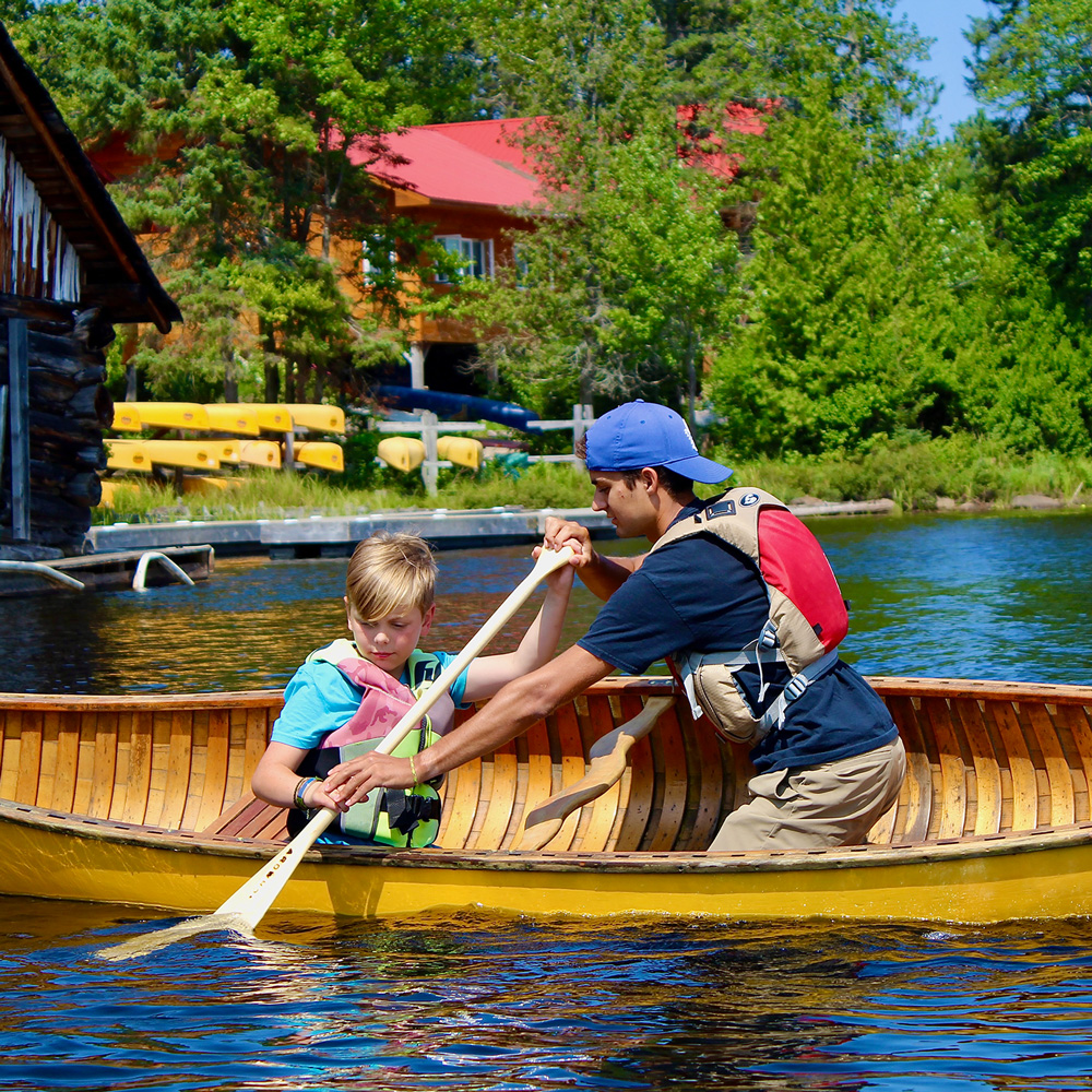 older camper helping younger camper paddle in the water at camp arowhon